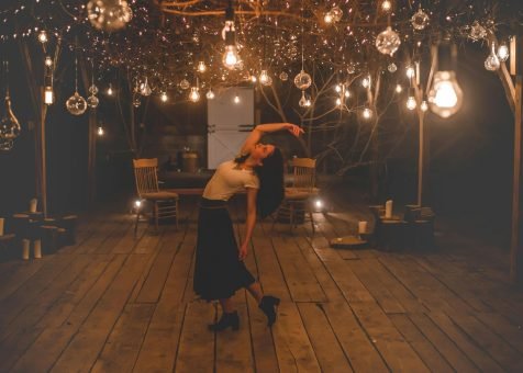 A woman gracefully dances under warm, illuminated bulbs in a rustic wooden interior.
